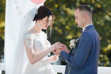 A bride and groom are getting married and the bride is wearing a white veil. The groom is wearing a blue suit and is about to give the bride a ring