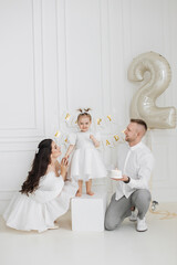 Caucasian family celebrating young girl's second birthday. Parents with two-year-old daughter in white dresses, smiling and holding cake.