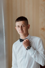 Wall Mural - A man in a white shirt and black bow tie is standing in front of a window. He is wearing a watch and he is getting ready for a formal event