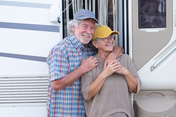 Cheerful senior couple on leisure trip standing outside a camper hugging each other and looking away. Beautiful Caucasian couple of retirees enjoying freedom and alternative life in motor home