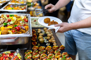 A person serving themselves a variety of dishes from a lavish buffet featuring gourmet appetizers and colorful fruits at a festive event