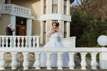 A bride is standing on a balcony of a large house, wearing a white dress. A man is standing behind her, holding a bouquet. The scene is likely a wedding or a special event