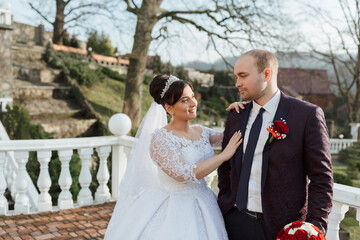 A bride and groom are standing on a balcony, with the bride wearing a white dress and the groom wearing a suit