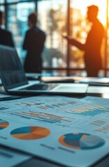 Poster - Close-up of business documents with graphs on a table in a bright office setting with silhouetted professionals in the background