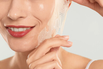 Poster - Smiling woman peeling off face mask on light grey background, closeup