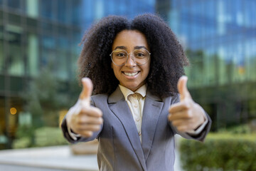 Confident African American business woman giving positive gesture outside office building. Professional attire, bright smile, symbolizing success, approval, motivation.