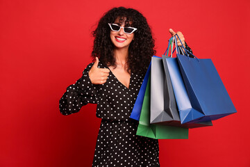 Smiling woman with colorful shopping bags showing thumbs up on red background