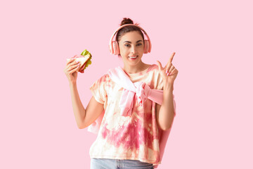 Canvas Print - Young woman in headphones with tasty sandwich pointing at something on pink background