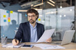 © Liubomir - Mature financier working diligently inside modern office reviewing paperwork with focus and precision using laptop and smartphone