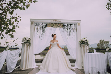 Wall Mural - A bride is standing in front of a white archway with a white canopy. She is wearing a white dress and is smiling. The scene is set in a garden, with a few potted plants and a bench nearby