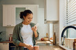 © Drazen - Happy Hispanic woman using cell phone while drinking coffee in kitchen.