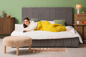 Poster - Beautiful young woman with warm yellow plaid and laptop lying on bed in bedroom