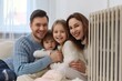 © miss irine - Happy family sitting near modern electric radiator at home. Adult, caucasian male, female with children, relaxing, smiling, feeling warmth in cozy room during autumn. Central heating system provides
