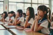 © miss irine - Classroom setting with students seated at desks in rows. Schoolgirls wear uniforms, engaged in lesson activity. Natural from window, whiteboard on wall. Students focus on teacher, notes, participate