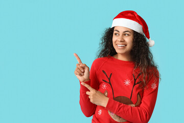 Poster - Happy young African-American woman in Christmas sweater and Santa hat pointing at something on blue background