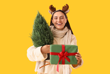 Wall Mural - Young woman in reindeer horns with small Christmas tree and gift box on yellow background