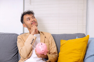 Young Asian Businessman Holding Piggy Banks While Thinking With Hand on Chin, Financial Planning and Investments Concept
