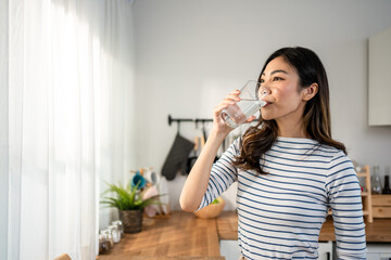 Wall Mural - Asian young woman drinking a glass of water in kitchen at home. 