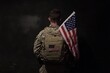 © adri - A soldier in camouflage uniform stands with his back to the camera, holding an American flag over his shoulder, against a dark background.