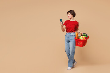 Wall Mural - Full body young happy woman wear red t-shirt casual clothes hold basket bag with food products use mobile cell phone isolated on plain light beige background. Delivery service from shop or restaurant.