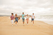 © Prostock-studio - A group of five friends run joyfully along the beach, laughing and embracing the warm atmosphere as waves crash nearby during a cloudy sunset.