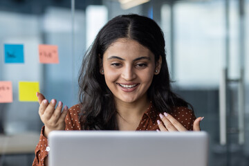 Wall Mural - Smiling woman communicates through laptop in contemporary office. Engaging in virtual meeting with gestures, surrounded by sticky notes. Represents technology, communication, remote work.