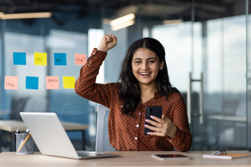 Wall Mural - Smiling young woman in office celebrating achievement. Holding phone and sitting at desk with laptop, expressing excitement. Colorful sticky suggest project planning or brainstorming session.