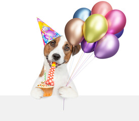Poster - Funny jack russell terrier puppy wearing party cap holding and cupcake and balloons, blowing in party horn and looking above blank white banner. isolated on white background