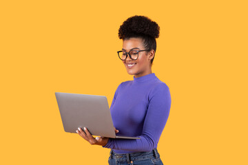 Poster - A young woman with curly hair and glasses smiles while using her laptop, standing in front of a vivid yellow background. Her cheerful demeanor suggests a productive moment in her work.