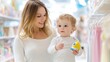 © Natalia - A 1-year-old Caucasian toddler is held by her smiling mother while they shop for clothes in a store