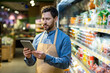 © Liubomir - Supermarket employee wearing apron uses tablet in grocery aisle. Man manages inventory with modern technology, demonstrating efficiency and focus in retail environment.