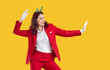 © Studio Romantic - Beautiful happy young brunette woman is fooling around and dancing funny, dressed in red business suit Santa Claus with figures of Christmas trees on her head. Model on isolated yellow background.