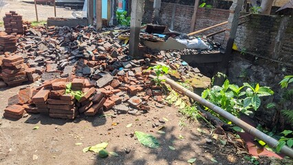 A large pile of bricks and rubble in front of a partially demolished building. A small temple with a blue facade and a brick wall are visible in the background. A pipe runs along the bottom.