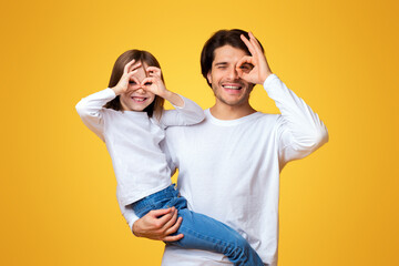 Funny little girl and daddy fooling, having fun over yellow background, imitating binoculars