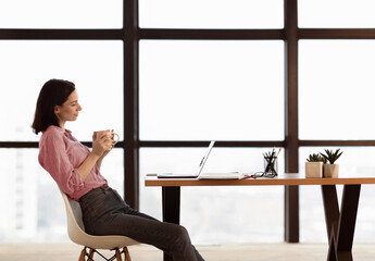 Poster - Coworking Concept. Side view of young female manager in shirt sitting at table, drinking coffee and working on laptop