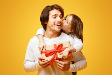 Wall Mural - Adorable little daughter and father holding gift box, girl kissing dad from behind, Father Day concept, yellow background