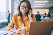 © BullRun - Portrait of successful female freelancer in casual spectacles for provide eyes protection sitting at desktop for distance working and making positive cellphone conversation during coffee break