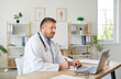 © Studio Romantic - Portrait of confident mature male doctor sitting at the desk and working in office looking at laptop monitor screen. Man physician in stethoscope in clinic making notes in notepad on workplace.