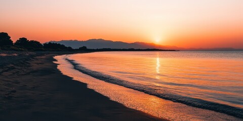 Wall Mural - Serene beach at sunset with soft waves, mountains in the distance, and a warm orange glow reflecting on the water.