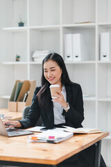 Wall Mural - professional woman in black suit is working on laptop while holding coffee cup, surrounded by office supplies and tidy workspace. Her focused expression conveys productivity and determination.