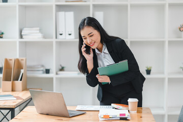 Wall Mural - professional woman in black suit is engaged in phone conversation while reviewing documents on her laptop. She appears focused and productive in modern office setting.