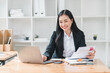 © kenchiro168 - professional woman is working at her desk, smiling while using laptop and organizing documents. bright office environment enhances her positive energy and productivity.