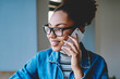 © BullRun - Positive female teenager in trendy spectacles for vision correction calling to friend for discussing time of meeting in cafeteria, happy hipster girl learning indoors and using mobile app for talking