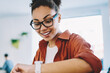 © BullRun - Positive smiling hipster girl in spectacles for provide eyes protection looking on modern smartwatch for manage time, cheerful african woman dressed in casual apparel checking notification