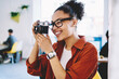 © BullRun - Cheerful female amateur photographer in optical spectacles making photo via vintage camera, dark skinned positive woman taking pictures using old fashioned equipment indoors enjoying time for hobby
