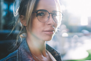 Young caucasian hipster girl in spectacles for provide eyes protection looking away during walking across town streets on sunny summer day, positive female teenager in eyewear spending time outdoors