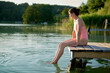 © Connect Images - Woman in a pink swimsuit sits on a wooden dock, dipping her feet in a tranquil lake during sunset.