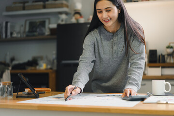 woman is working on project at wooden table, using pen to write on large sheet of paper. She appears focused and engaged in her task, surrounded by cozy home environment.