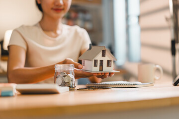 woman holds miniature house model while sitting at desk filled with coins, notebook, and cup. This scene reflects concept of home ownership and financial planning.