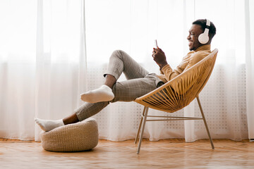 Aufkleber - Weekend. Cheerful Afro Man In Wireless Earphones Using Cellphone Listening To Music Sitting On Modern Chair Against Window At Home